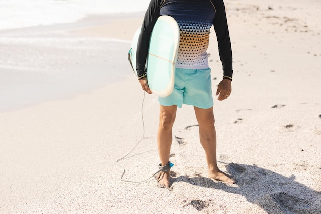 Photo low section of biracial senior man holding surfboard standing on shore at sunny beach