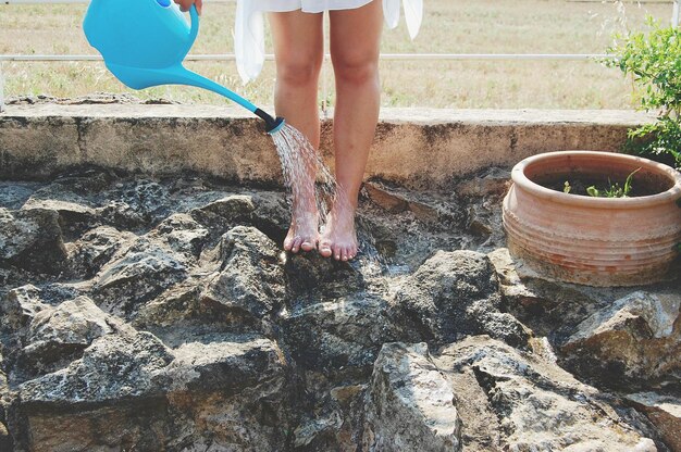 Foto sezione bassa di una ragazza che si lava i piedi con una lattina d'acqua sulle rocce all'aperto