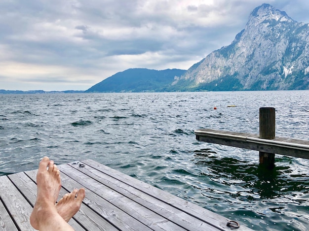 Low section of man relaxing on pier at sea against mountains