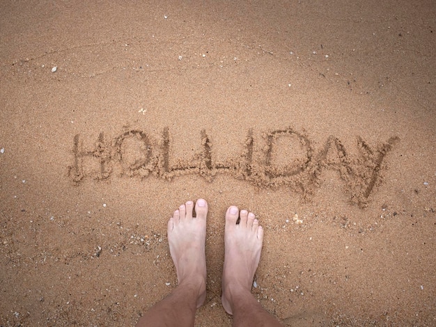 low section of man standing by holiday text in sand on shore at beach