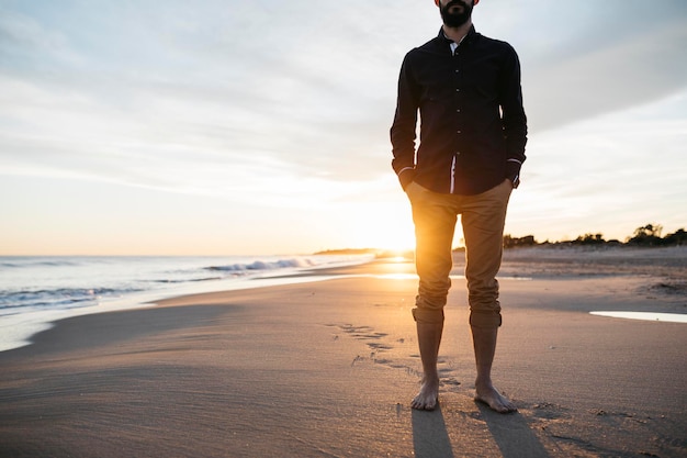 low section of mid adult man standing at beach against sky during sunset
