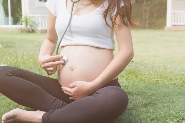 Foto sezione inferiore di una donna incinta che tiene uno stetoscopio sopra l'addome mentre è seduta sull'erba