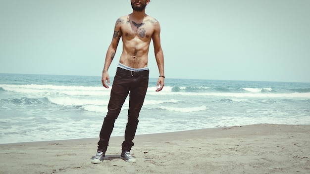 Low section of shirtless man with tattoos standing at beach