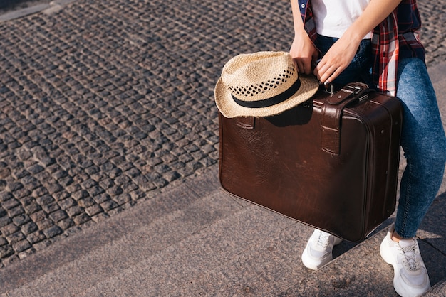 Foto sezione bassa della donna che porta la borsa per bagagli marrone con cappello in piedi sulla scala