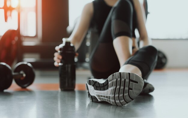 Foto sezione bassa di una donna che tiene una bottiglia d'acqua mentre è seduta sul pavimento in palestra