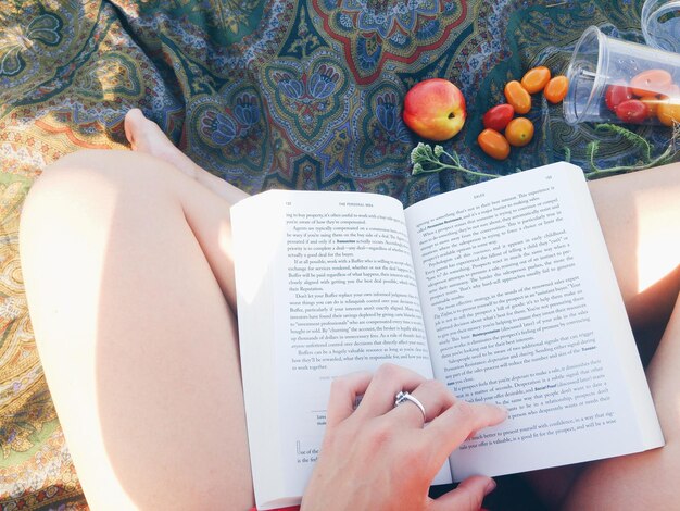 Photo low section of woman reading book by fruits on blanket during sunny day
