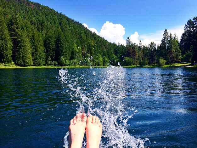 Low section of woman splashing river water against trees at forest