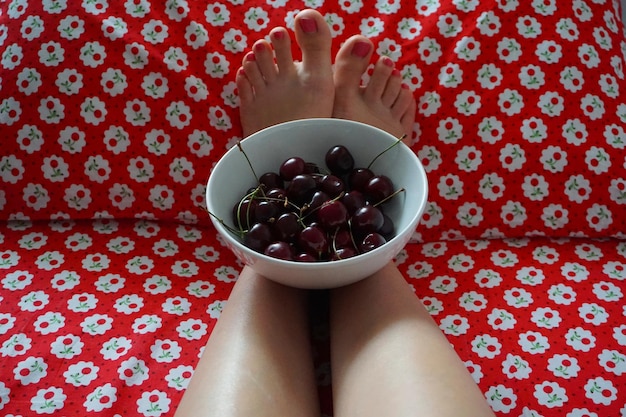 Foto sezione bassa della donna con le ciliegie in ciotola seduta a casa