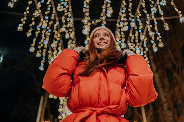 Lowangle beeld van een gelukkige jonge vrouw in een warme hoed en winterjas die op een straat in de sneeuwstad poseert