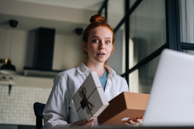 Lowangle shot van geschokte roodharige jonge vrouw die geschenkdoos opent met heden tijdens videogesprek op laptop zittend aan bureau Concept van vrijetijdsbesteding roodharige vrouw thuis tijdens zelfisolatie