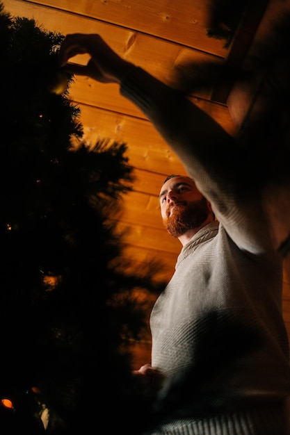 Lowangle vertical shot of young man in cozy sweater decorating Christmas tree with colorful shiny balls in dark living room