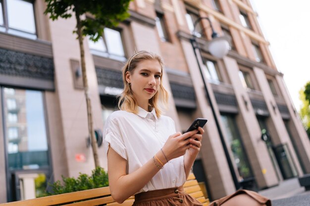 街の通りのベンチに座り、スマートフォンを手に持ったカメラを見ている魅力的な女性の金髪の若い女性のローアングル ビュー