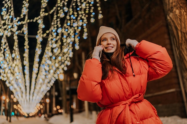 Lowangle view of happy young woman in hat and winter jacket talking using mobile phone standing on