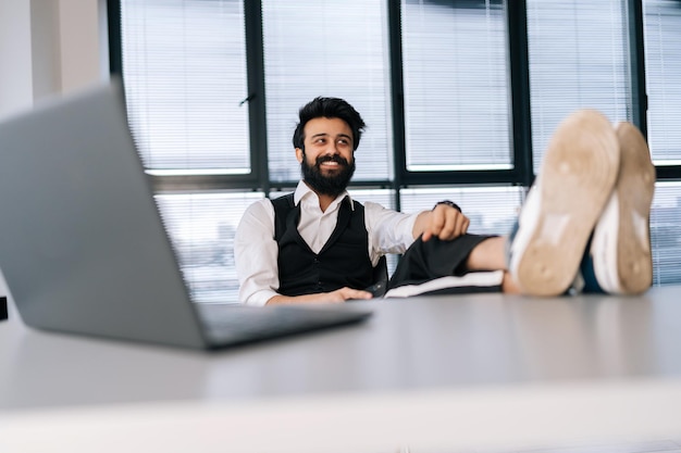 Lowangle view portrait of cheerful businessman sitting at office desk with feet up on table with laptop finished work job done completed all tasks relaxing after workday smiling looking away