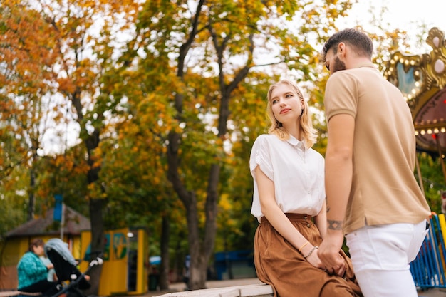 Низкий угол обзора романтической молодой влюбленной пары, стоящей, держащейся за руки, в парке развлечений на фоне карусели в летний день
