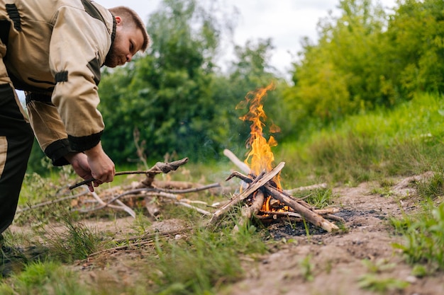 Lowangle view of skilled fisher man putting firewood on campfire to making fire on bank of river before cooking in evening before sunset