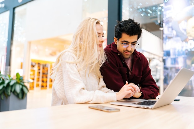 Lowangle view of young couple using typing laptop computer\
together discussing purchase on the internet looking to screen\
sitting at table in cafe