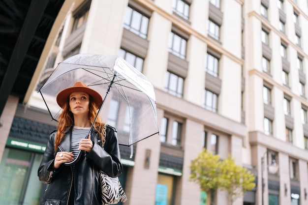 Lowangle weergave van charmante roodharige jonge vrouw in mode hoed wachtend in stadsstraat met transparante paraplu in regenachtig weer regen wegkijkend