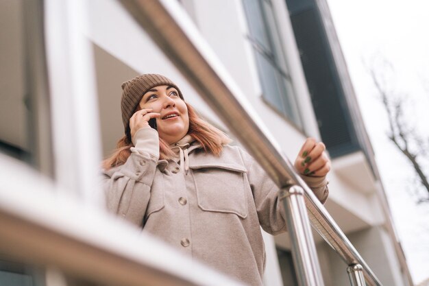 Lowangle weergave van overgewicht vrouw in warme muts en jas praten op smartphone staande in de buurt van reling op stadsstraat in bewolkte herfstdag