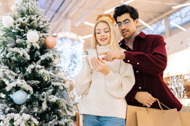 Lowangle weergave van vrolijk jong stel met behulp van mobiele telefoon samen staande met papieren zakken in de hal van het winkelcentrum vieren op kerstavond
