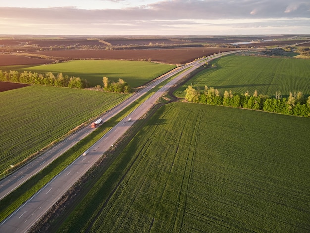 Luchtfoto drone weergave van een vrachtwagens rijden op asfaltweg langs de groene velden in het landelijke landschap bij zonsondergang.