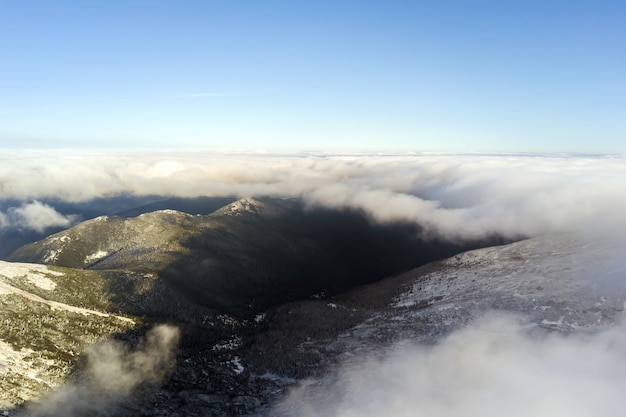 Luchtfoto van bovenaf van witte gezwollen wolken die besneeuwde bergtoppen bedekken in heldere zonnige dag.