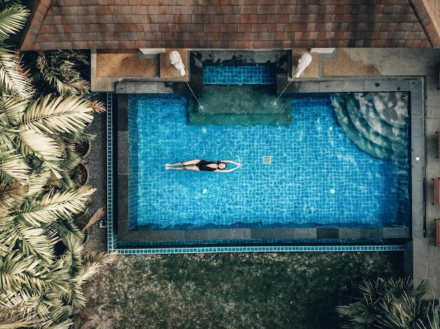 Luchtfoto van het hotelgebied met palmen, groen gazon, pannendak en het heldere zwembad met een slank meisje dat op de rug drijft; luxe concept.