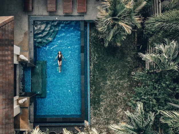 Luchtfoto van het hotelgebied met palmen, groen gazon, pannendak en het heldere zwembad met een slank meisje dat op de rug drijft; luxe concept.