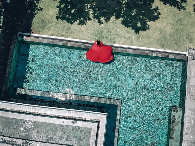 Luchtfoto van het prachtige meisje in het zwembad van het hotel. De jonge dame draagt een rode jurk die in het water stroomt; mode-concept.