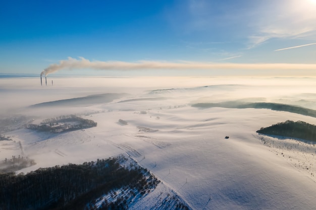 Luchtfoto van het winterlandschap met mistig landschap en verre fabriekspijpen die zwarte, vuile rook uitstoten die de omgeving vervuilt.