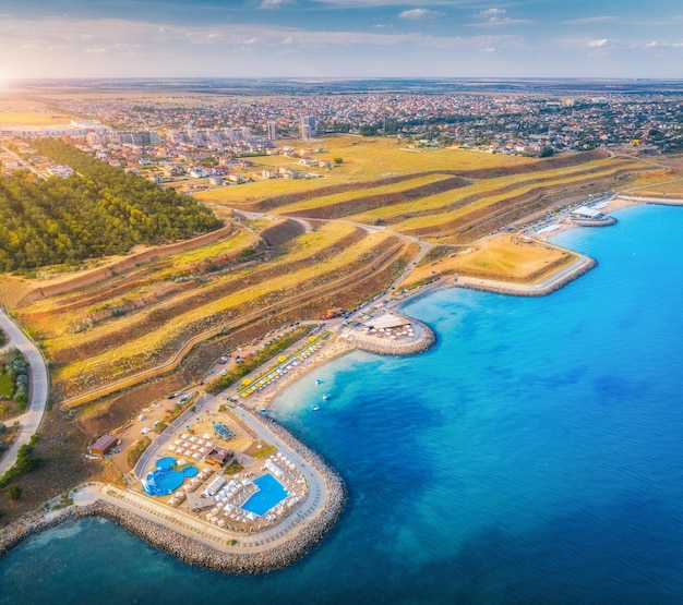 Luchtfoto van prachtig zandstrand blauwe zee restaurants aan de promenade zwembad parasols zwemmen mensen in helder water groene bomen bij zonsondergang in de zomer bovenaanzicht van de stad gele heuvels aan de kust