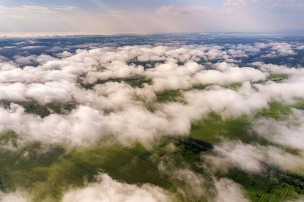 Luchtfoto van witte wolken boven een stad of dorp met rijen gebouwen en bochtige straten tussen groene velden in de zomer. Platteland landschap van bovenaf.