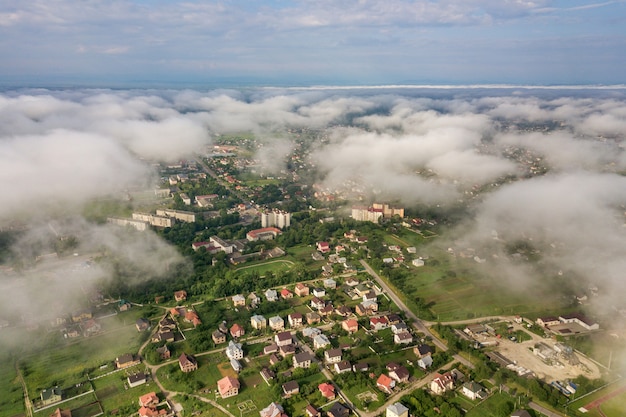 Luchtfoto van witte wolken boven een stad of dorp met rijen gebouwen en bochtige straten tussen groene velden in de zomer. Platteland landschap van bovenaf.