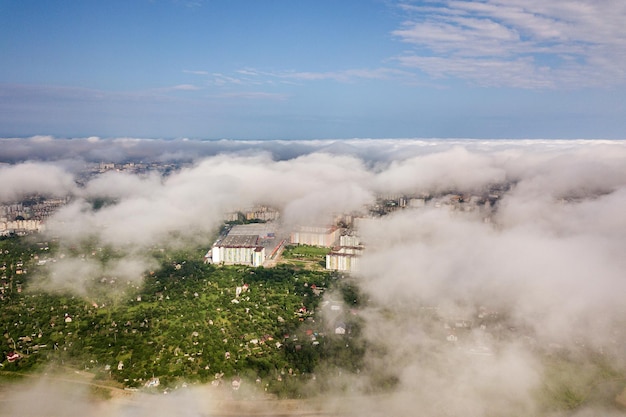 Luchtfoto van witte wolken boven een stad of dorp met rijen gebouwen en bochtige straten tussen groene velden in de zomer plattelandslandschap van bovenaf