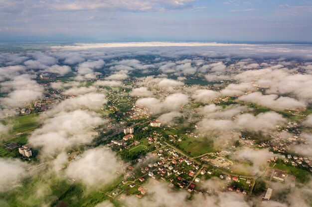 Luchtfoto van witte wolken boven een stad of dorp met rijen gebouwen en bochtige straten tussen groene velden in de zomer.