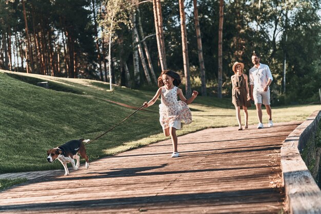 Lucky to be together. Full length of cute little girl running with dog and smiling while her parents walking behind