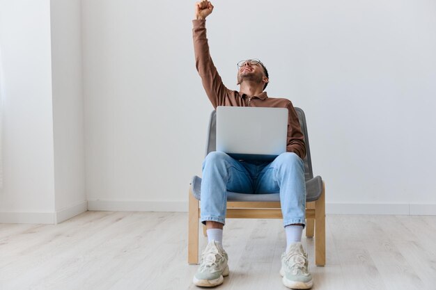 Lucky cheerful overjoyed young tanned man with laptop on knees\
raise fists up celebrating big win say yeah sitting on chair at\
white home wall lottery winner offer copy space