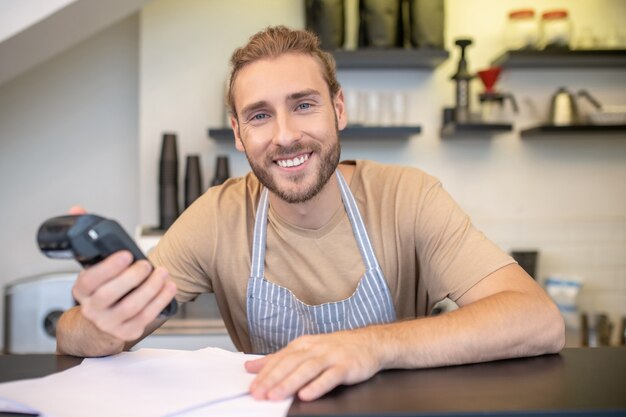 Lucky day. Happy smiling young bearded man with pos terminal in hand at bar counter checking bills in cafe