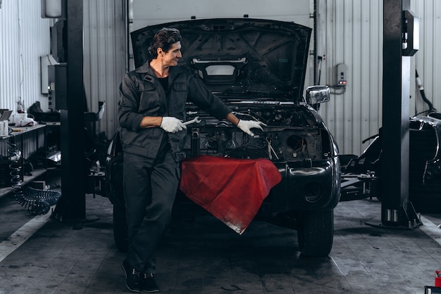 幸運な日。大きな車のガレージで黒い開いた車の近くに立っている間、作業服のポーズで大人の男性の笑顔