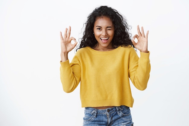 Lucky goodlooking african american woman in yellow sweater wink and show affirmative gesture okay signs approve amazing good concept satisfied with awesome product white background