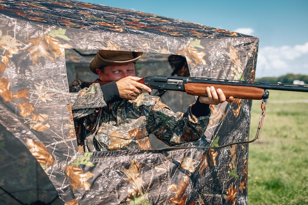 Lucky hunter sitting in wait, the man holds a rifle standing in a camouflage tent wearing a khaki hat; hunting concept.