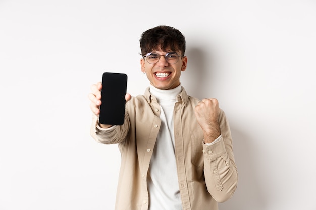 Lucky young man winning prize online, showing smartphone screen and rejoicing of good news, standing happy on white wall.