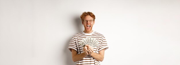 Photo lucky young man with red hair showing dollars winning money and screaming of happiness holding prize