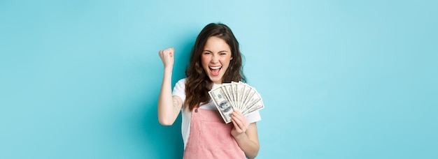 Photo lucky young woman looks excited shouting from satisfaction and triumph winning money holding dollar bills and making fist pump standing over blue background