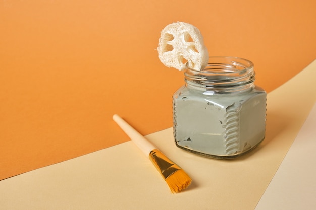 Luffa, brush, cosmetic clay in a glass jar on a wooden stand, beige and brown background, copy space