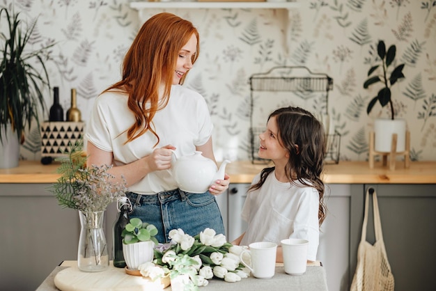 Luier mooie aanhankelijke moeder met een boeket lentebloemen schenkt thee aan haar dochter knuffelt haar dochter thuis in de keuken geniet van het moment van verrassing viert Moederdag