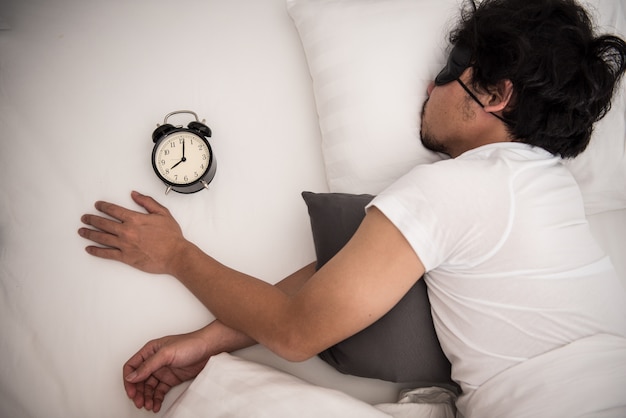 Luiheid van de Aziatische mens die nog op het bed in slaapkamer in ochtend met zwarte wekker slaapt.