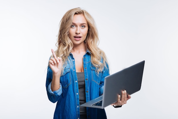 Luister eens. Mooie blonde vrouw die een laptop vasthoudt en haar vinger opheft, een belangrijke opmerking maakt terwijl ze geïsoleerd op een grijze achtergrond staat