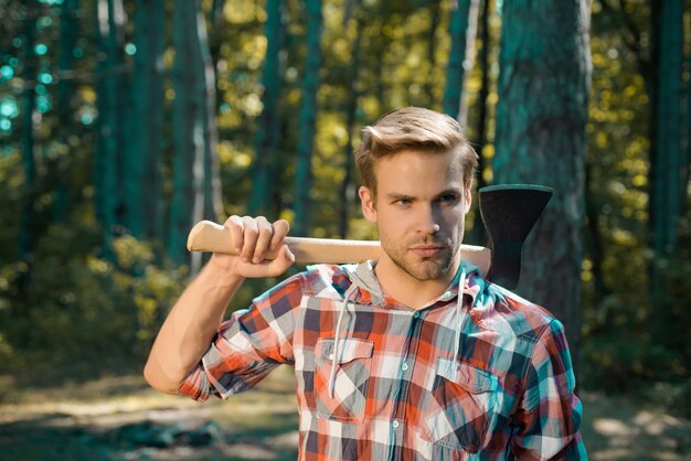 Lumberjack on serious face carries axe on shoulder Bearded man with axe concept Lumberjack with axe on forest background Man doing mans job Harvest of timber