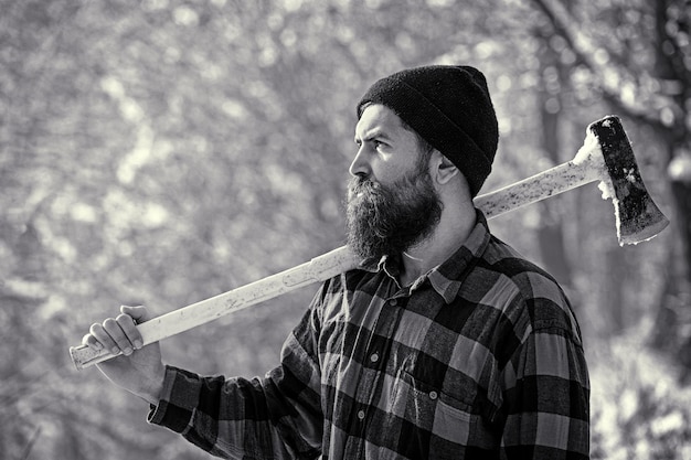 Lumberjack in the woods with an ax. Bearded man in hat with a hatchet. Handsome man, hipster. Lumberjack brutal bearded man with beard and moustache on winter day, snowy forest. Black and white.